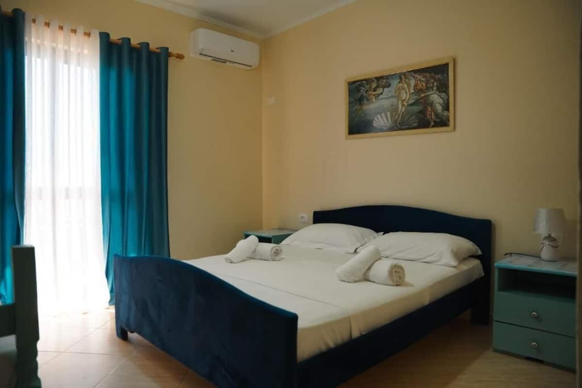 A bedroom with a bed, a nightstand, a painting on the wall, and a window with curtains.