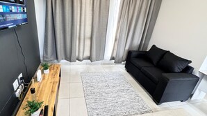 A modern living room with a black sofa, a wooden coffee table with plants, a grey rug, and a TV mounted on the wall.