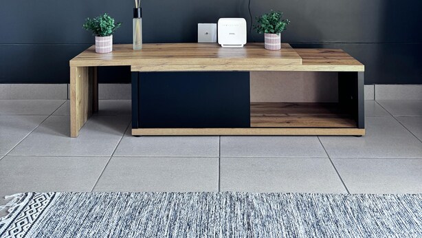 A modern living room with a flat-screen TV mounted on a dark wall, a wooden bench with a built-in white speaker, and two potted plants.