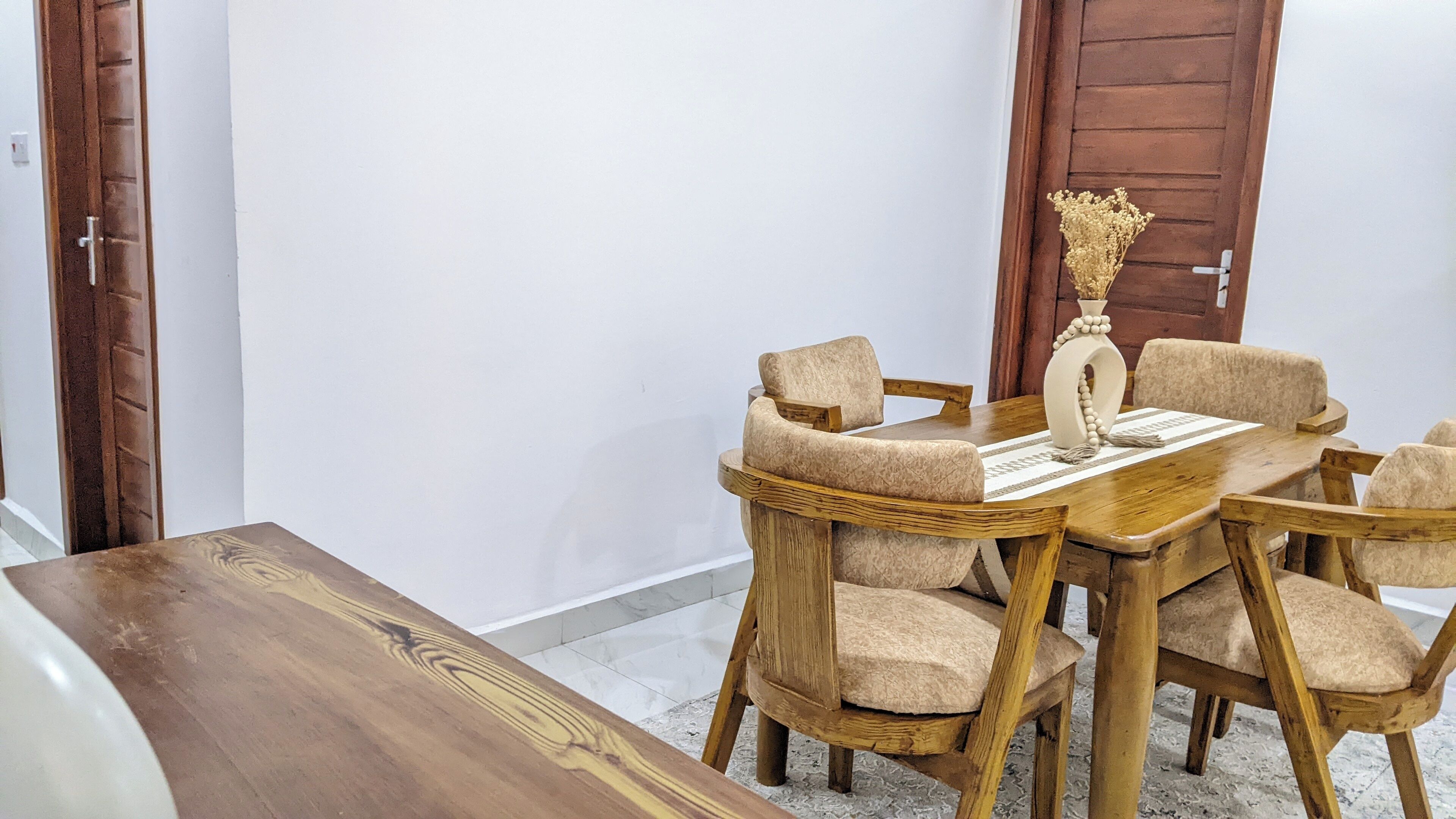 A dining area with a wooden table and chairs, a vase with dried flowers, and a door in the background.