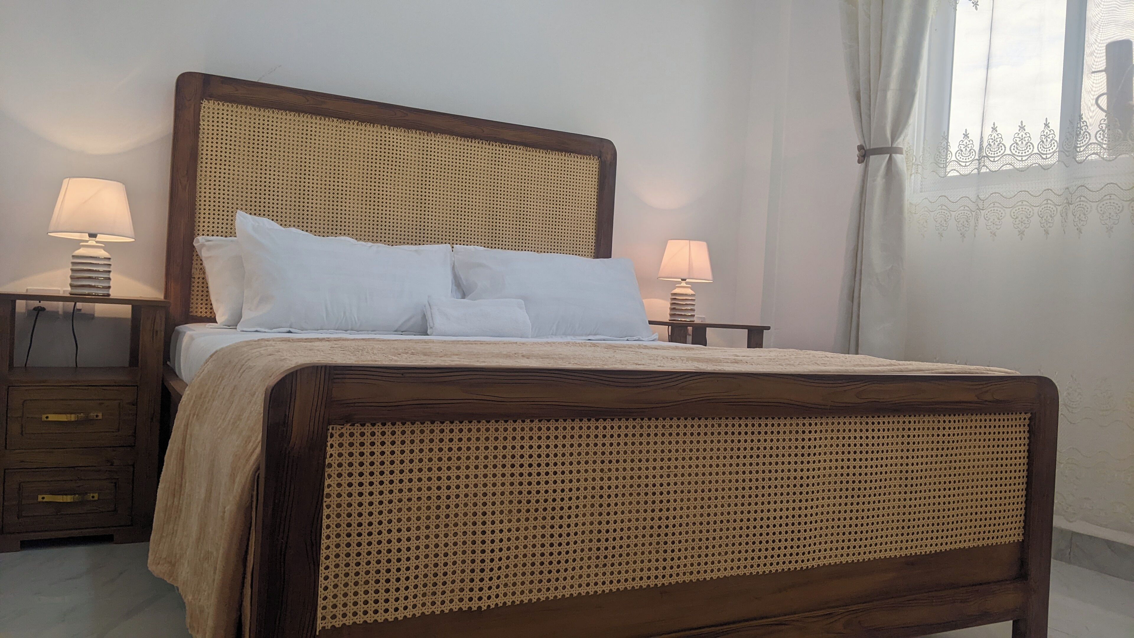 A wooden bed with a woven headboard, two bedside tables with lamps, and a window with curtains.