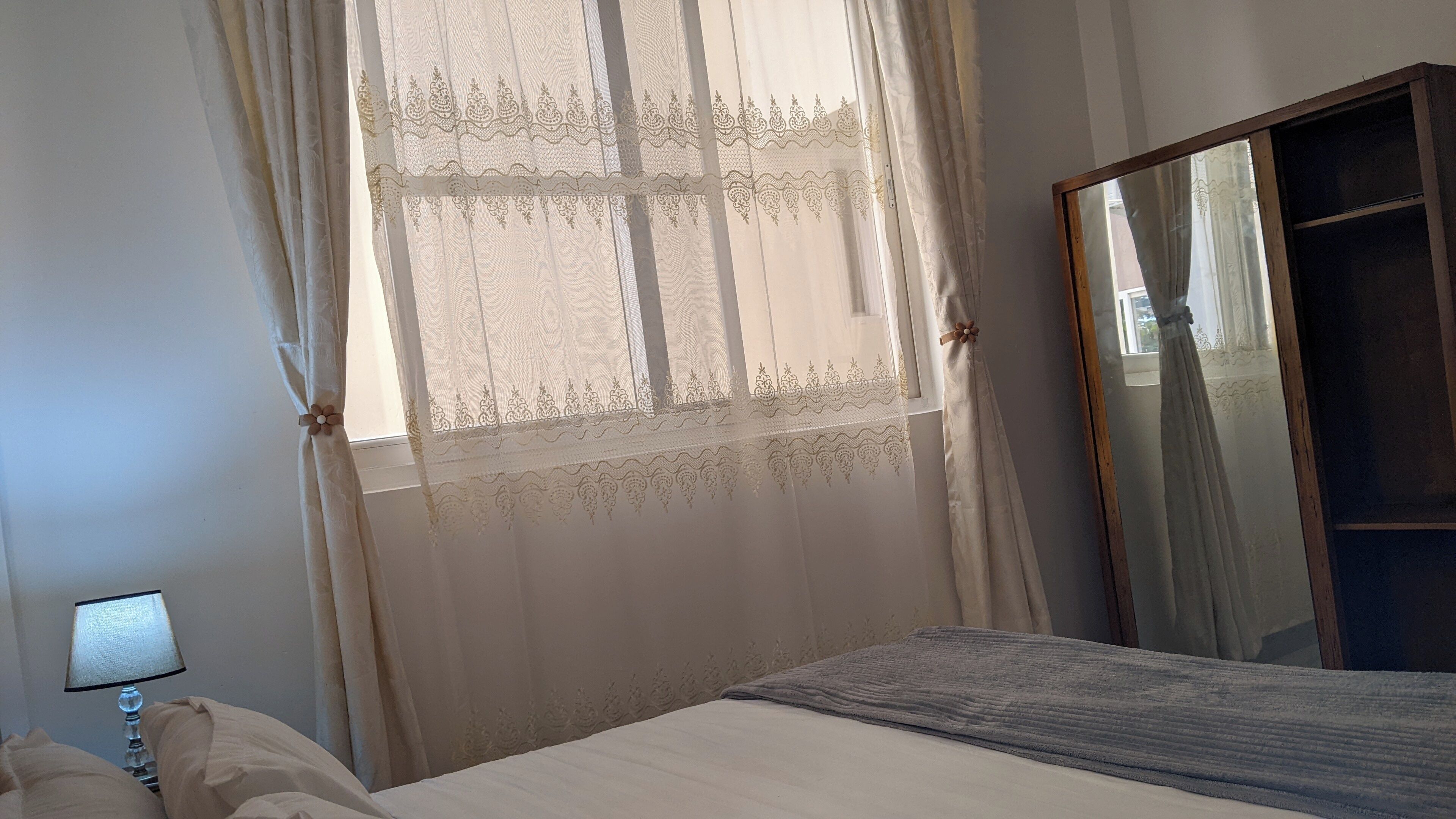 A bedroom with a bed, a nightstand, a lamp, a window with sheer curtains, and a mirror.