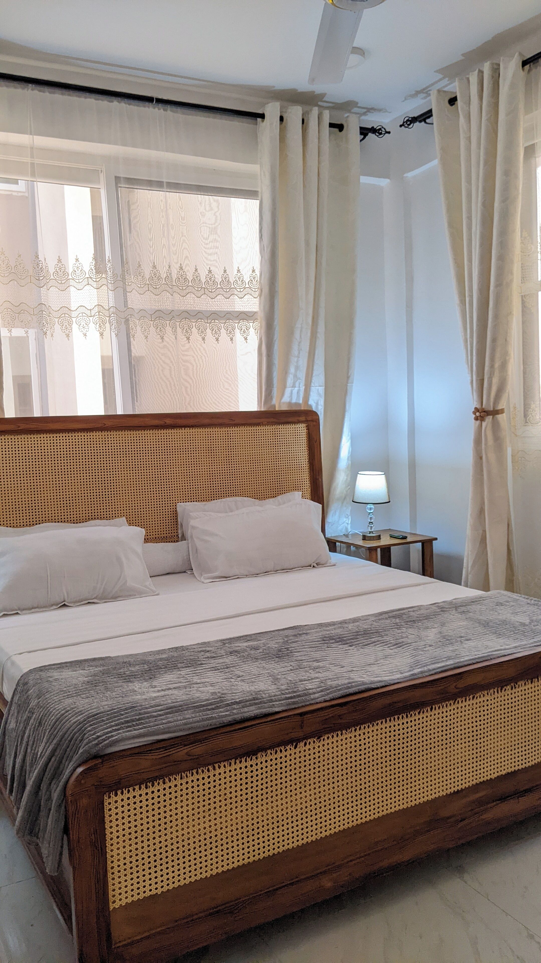 A bed with a woven headboard, a bedside table with a lamp, and sheer curtains.