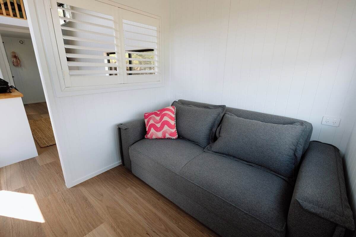 A grey sofa with a pink and grey pillow, a white window with blinds, and a wooden floor.