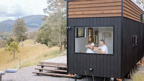 Une petite cabane moderne, avec une partie supérieure en bois et une partie inférieure de couleur foncée. Elle est dotée d’une grande fenêtre et d’une terrasse en bois.