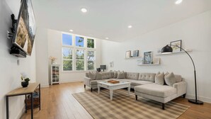 A living room with a sectional sofa, a coffee table, a TV mounted on the wall, and a window with blinds.