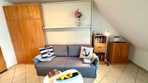 Una sala de estar acogedora con un sofá gris, una mesita blanca con frutero, armarios de madera y una estantería.