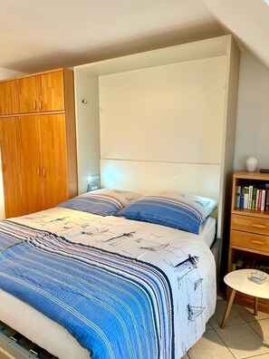 Una camera da letto con un letto, un armadio in legno, un comodino con una lampada e un tavolino rotondo con dei libri.