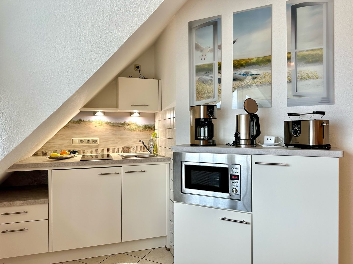 Una cocina moderna con armarios blancos, horno empotrado, microondas y cafetera. Dos cuadros enmarcados están sobre la encimera.