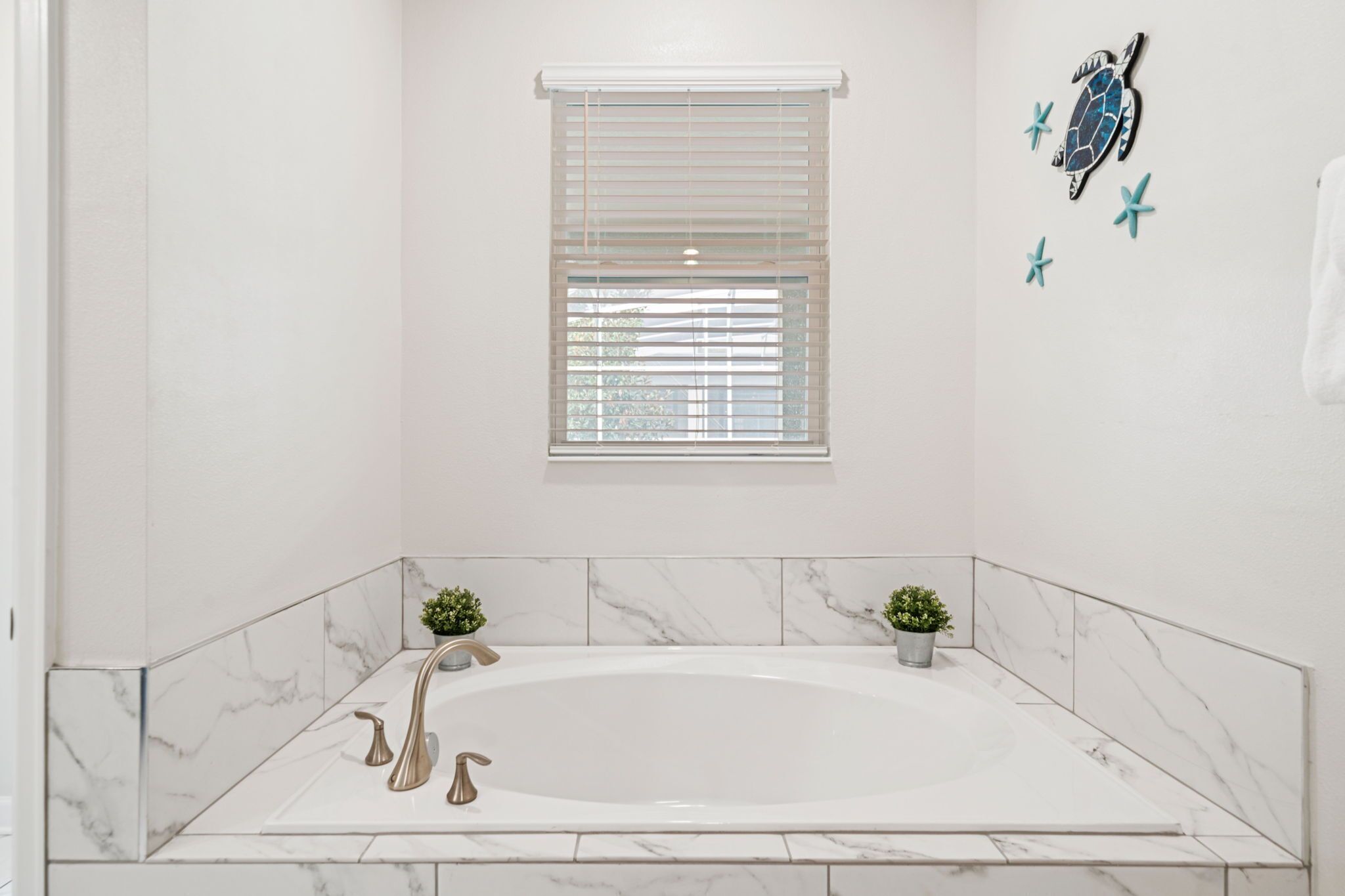Une salle de bain dotée d’une grande baignoire, de murs en marbre blanc, d’une fenêtre avec stores et de décorations murales représentant des tortues de mer et des étoiles de mer.