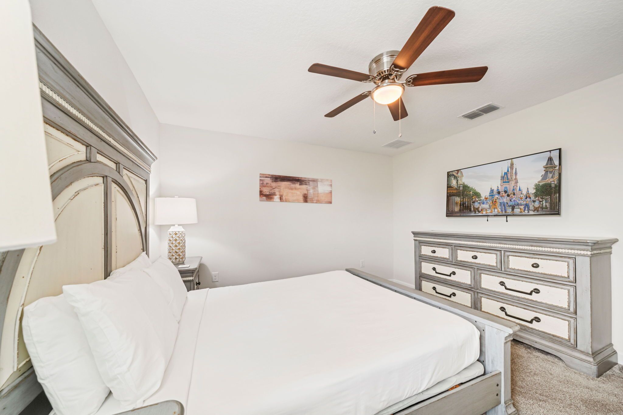 Une chambre à coucher comprenant un lit, un ventilateur de plafond, une commode et un tableau accroché au mur.