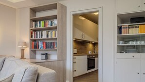 Uma sala de estar moderna com um sofá, uma estante e uma cozinha visível através de uma porta aberta.