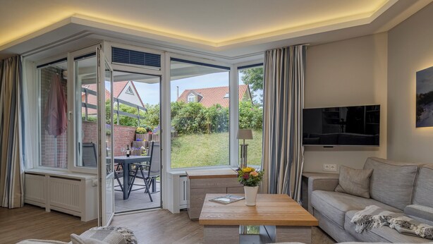 Uma sala de estar moderna com sofá, mesa de centro e televisão. Há uma janela grande com cortinas, vista para o jardim e uma área de jantar visível.