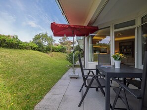 An outdoor patio with a table and chairs under a red umbrella, a glass door leading to a living area, and a grassy yard. - Modern vacation apartment with 3 rooms on approx. 87 m², for max. 4 persons (Westerland)
