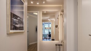Um corredor com um quadro na parede à esquerda, uma porta que dá para um quarto com janela, e uma mesinha com abajur à direita.