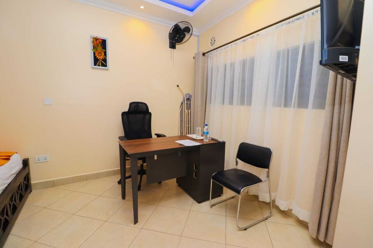 Habitación de hotel con escritorio, dos sillas, cama, televisor, ventilador y ventana con cortinas.