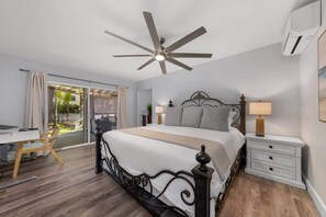 A bedroom with a bed, a desk, a chair, a ceiling fan, a nightstand, and an air conditioning unit.