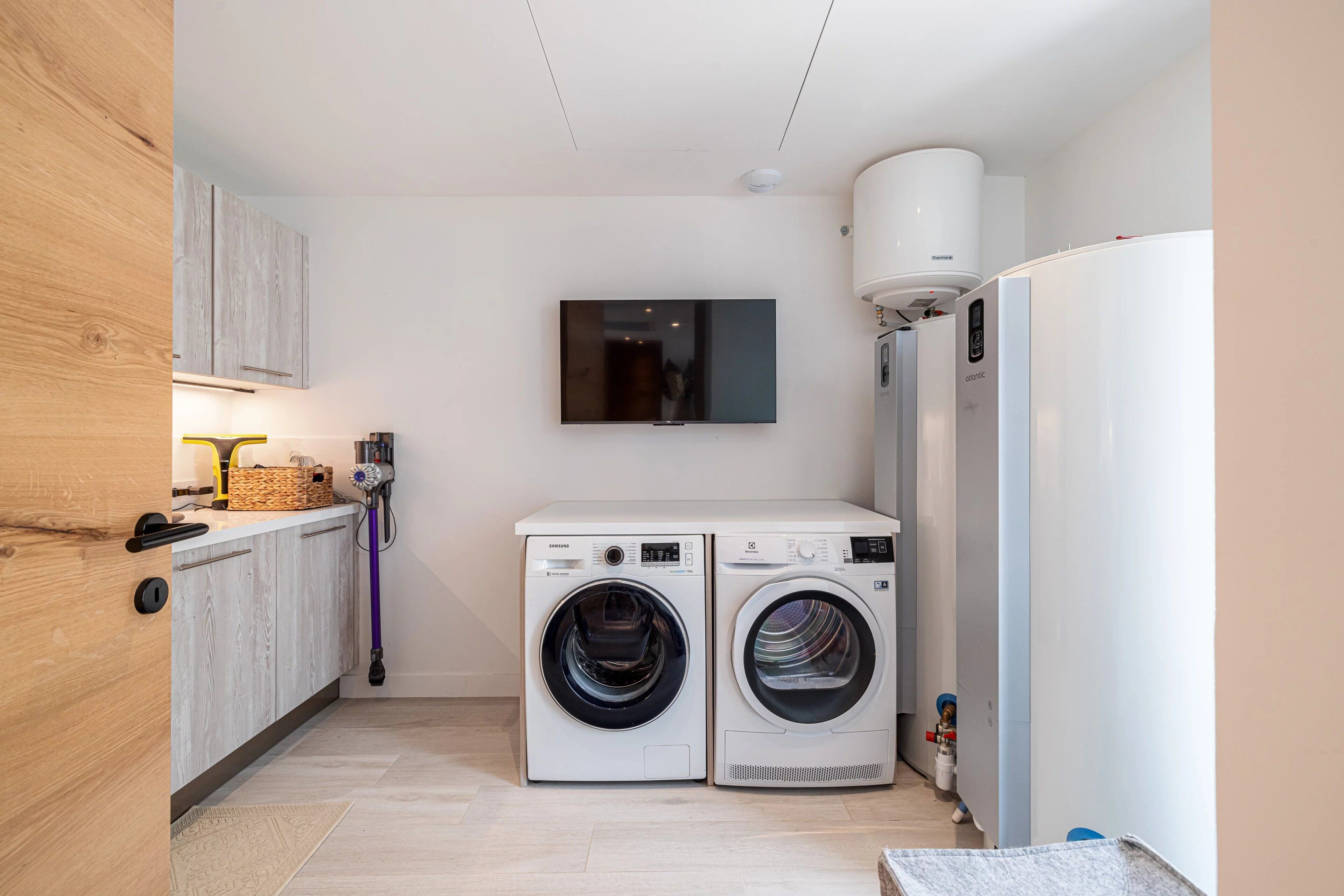 A compact laundry room with two front-loading washing machines, a wall-mounted television, and a built-in refrigerator.
