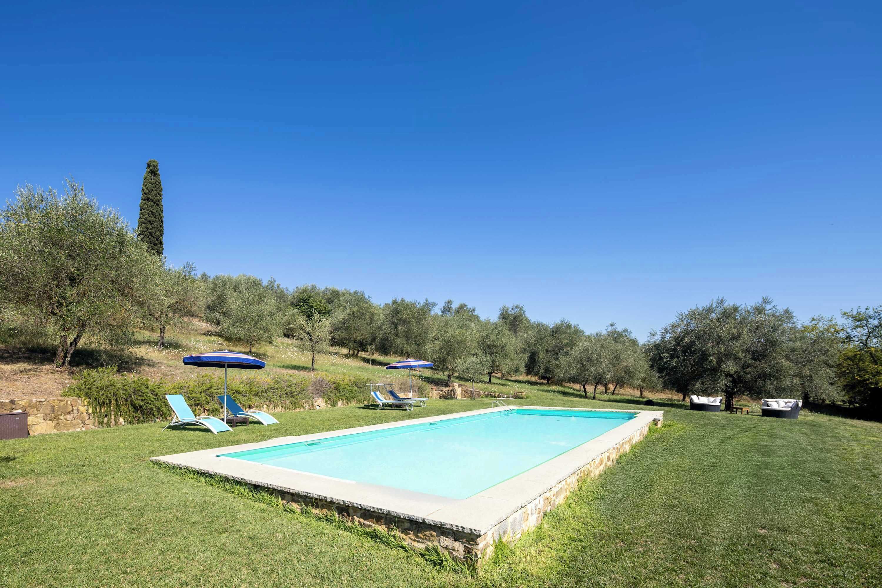 Une piscine rectangulaire bordée de pierres, entourée d’une pelouse bien entretenue. On y trouve deux chaises longues et un parasol bleu, près de la piscine.
