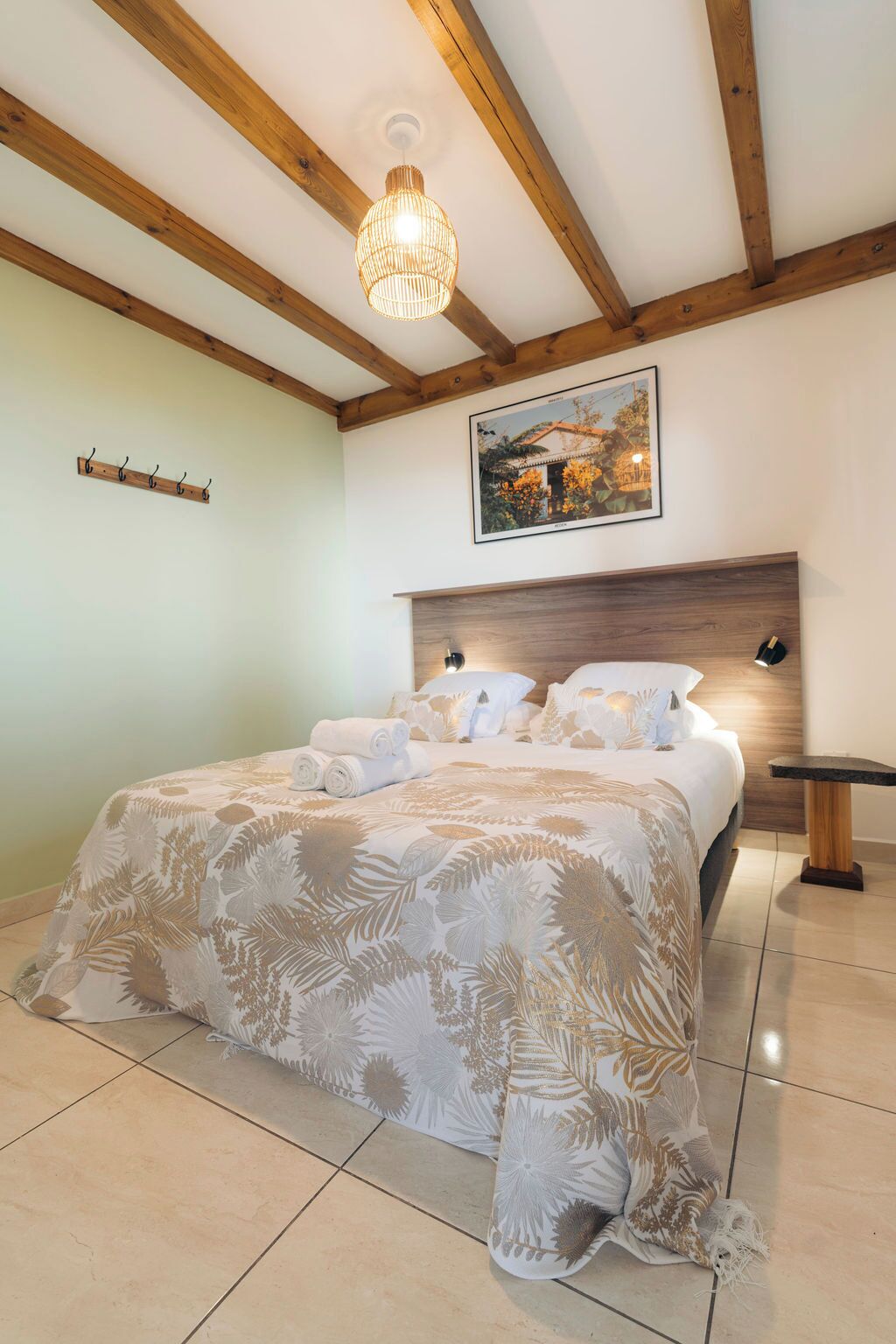 A bedroom with a bed, wooden beams, a hanging light fixture, a wall-mounted picture, and a small table.