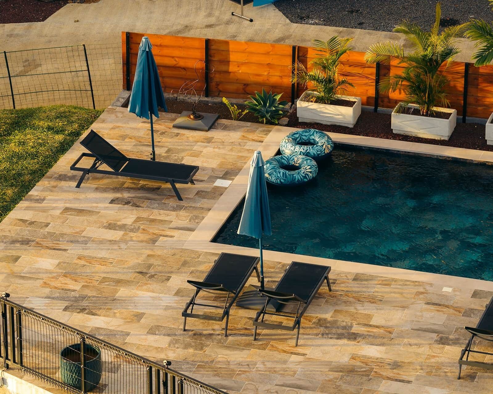 Une piscine extérieure entourée d’une clôture en bois et agrémentée de plantes en pot, avec des chaises longues et des parasols à proximité.