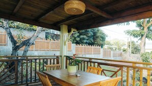 Een houten terras met een tafel en stoelen, een hanglamp en uitzicht op een omheinde tuin met bomen.