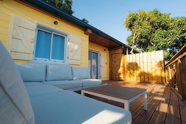 Una terraza de madera con un sofá blanco y una mesa, un cerco de madera y un edificio amarillo con contraventanas.