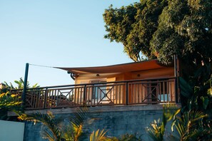 Una terraza de madera con barandilla, con vista a un edificio y un árbol grande al fondo.