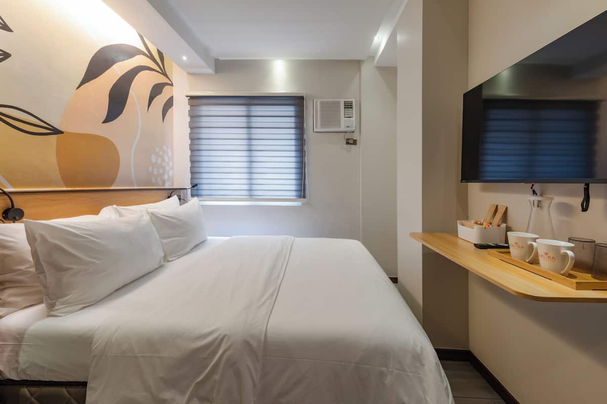 A hotel room with a bed, a wall-mounted TV, a window with blinds, a small table with cups and a tray, and a wall decoration.