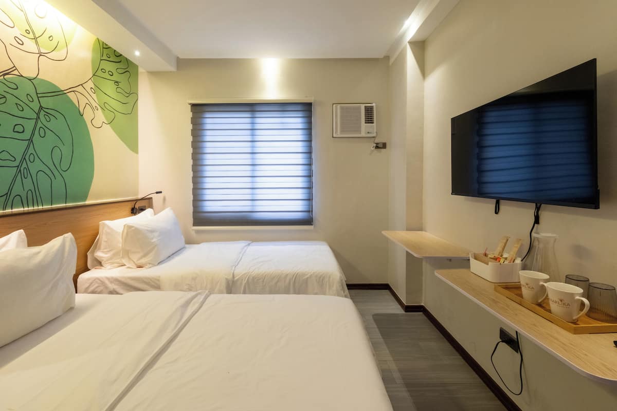 A hotel room with two beds, a flat-screen TV, a window with blinds, and a wall-mounted air conditioner.