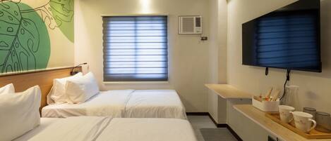 A hotel room with two beds, a flat-screen TV, a window with blinds, and a wall-mounted air conditioner.