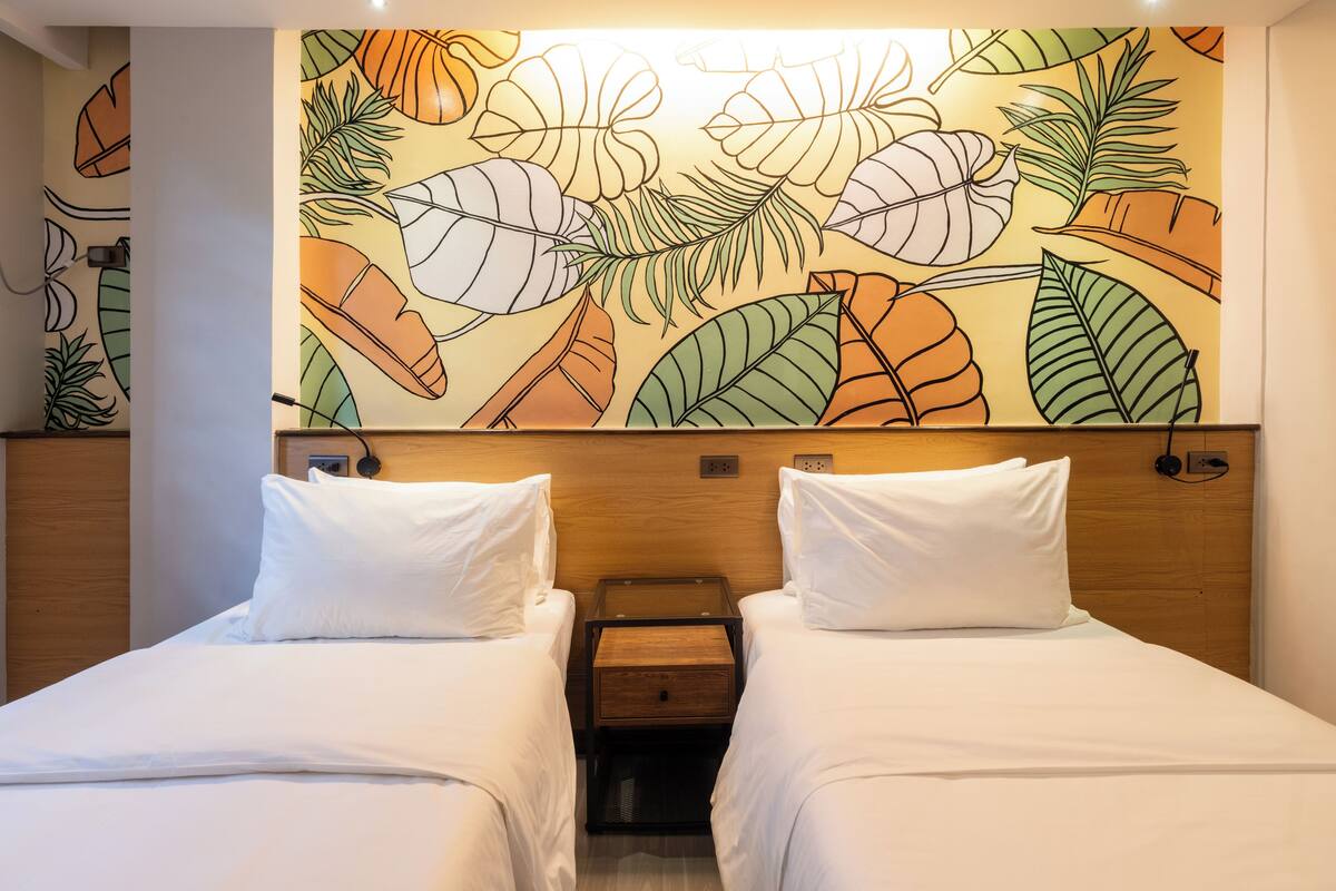A hotel room with two beds, a wooden headboard, and a wall mural of tropical leaves.