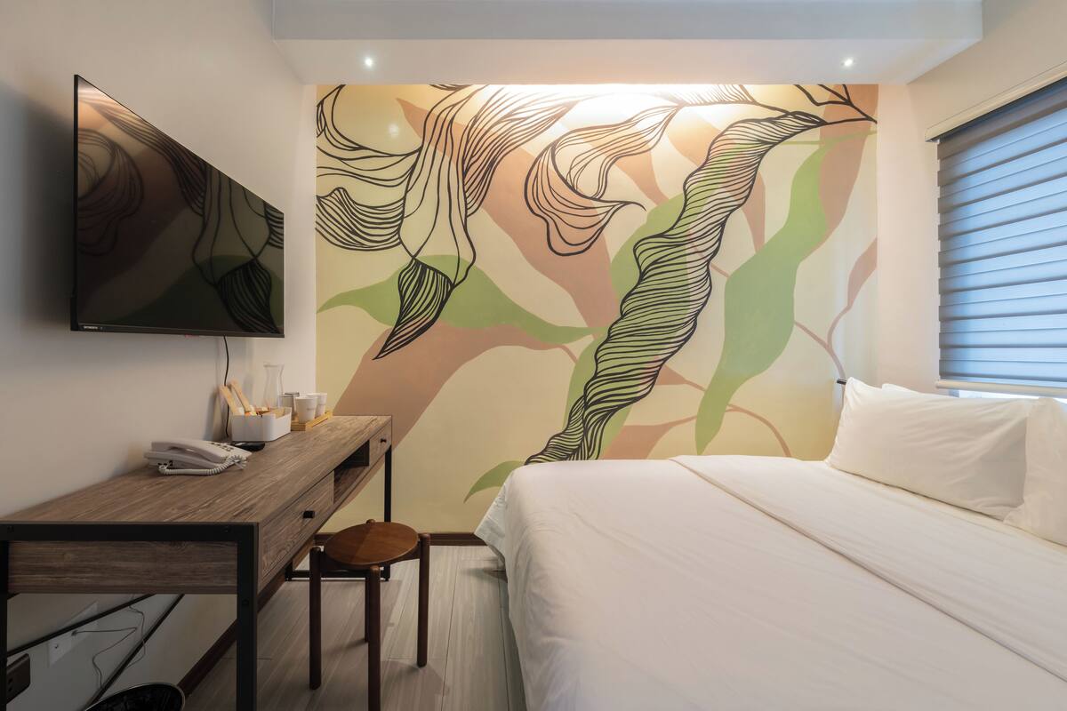 A hotel room with a bed, a desk with a telephone, a chair, and a television mounted on the wall.