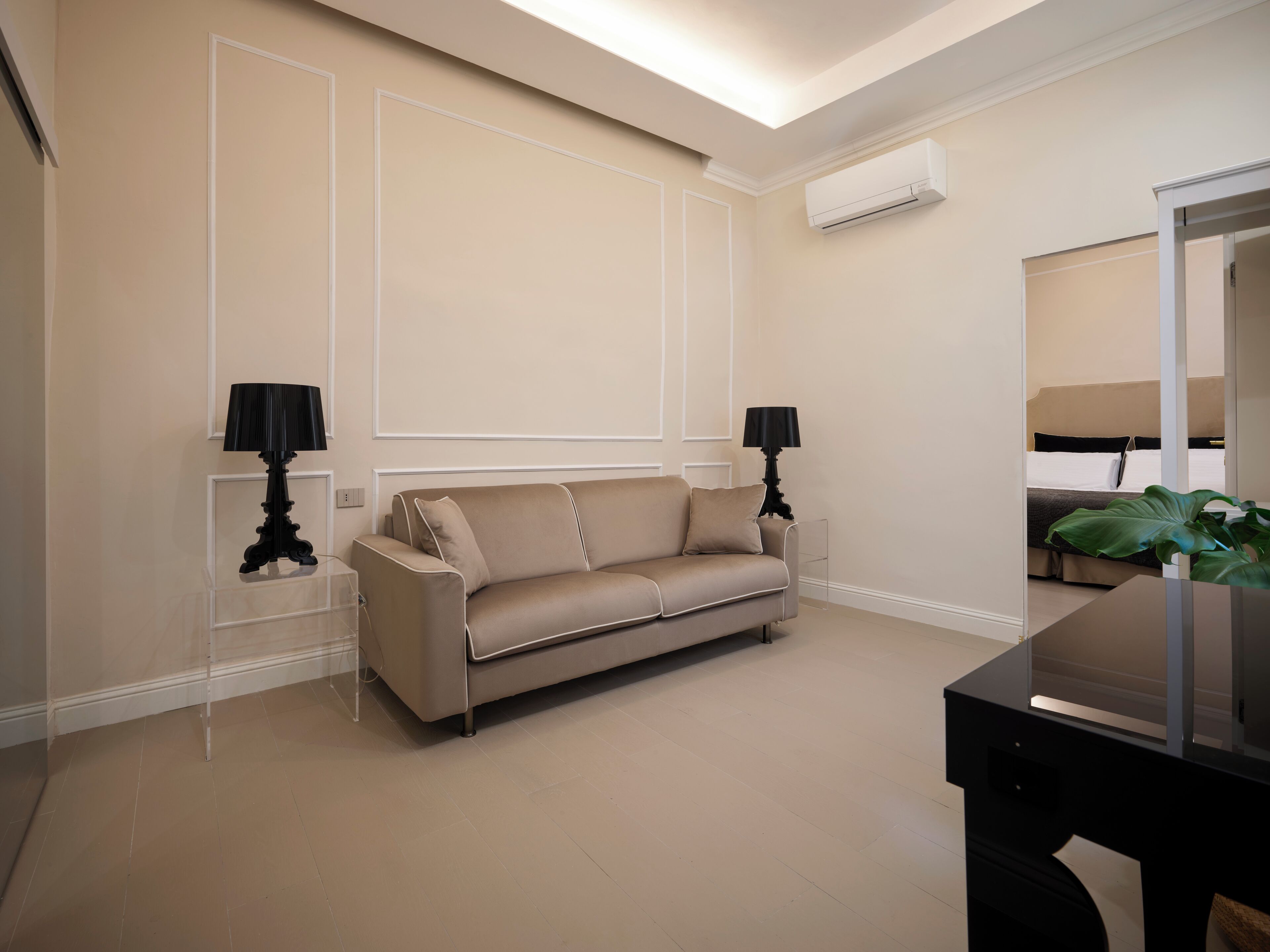 Quarto de hotel moderno com sofá bege, duas luminárias pretas, um espelho grande e um aparelho de ar condicionado na parede.