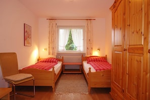 Une chambre à coucher avec deux lits en bois, un radiateur, une fenêtre avec des rideaux, une chaise et une armoire.