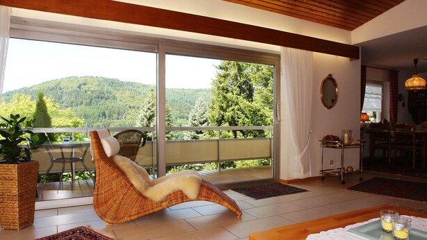 Une pièce avec un fauteuil en osier, une table à manger avec un set de table, et une vue sur une colline boisée, visible à travers de grandes portes-fenêtres coulissantes.