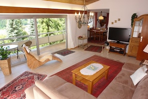 A living room with a sofa, a coffee table, a TV, a dining area, and a balcony with a view.