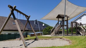 Une aire de jeux avec des balançoires et une structure d’escalade, un espace abrité et, au fond, un bâtiment doté d’un toit en pente.