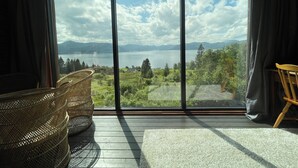 A room with a large sliding glass door leading to a scenic view of a lake and mountains. There are wicker chairs and a wooden chair inside the room.