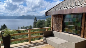 A wooden deck with a wicker sofa and a potted plant, overlooking a lake and mountains.