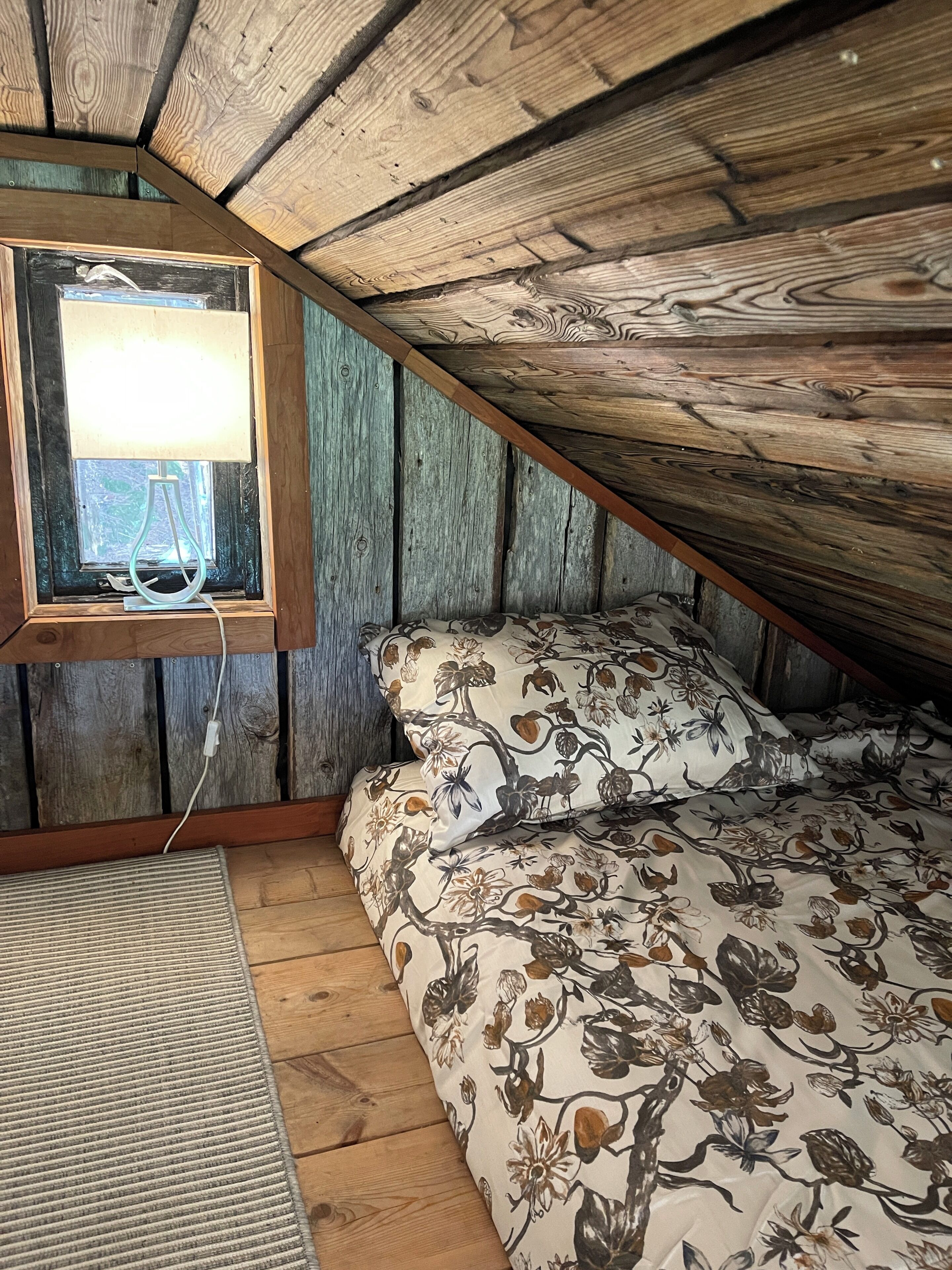 A cozy attic bedroom with a sloped ceiling, wooden walls, a window with a lamp, floral bedding, and a rug.