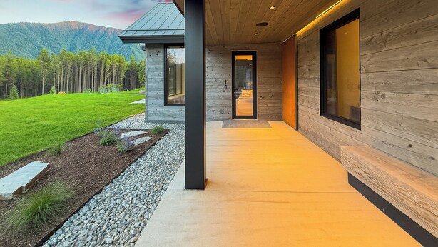A modern building with a wooden exterior, a concrete pathway, and a view of a green landscape with trees and mountains in the background.