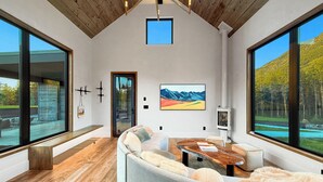 A modern living room with a large sectional sofa, a wooden coffee table, and a fireplace. There are large windows offering a view of the outdoors.