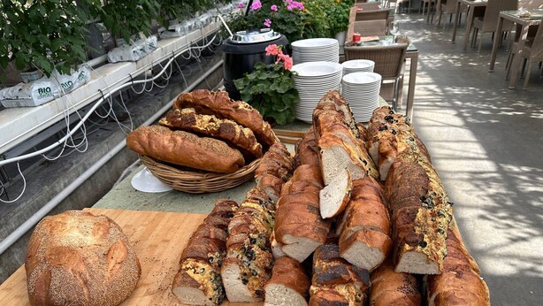 A variety of breads on a wooden cutting board, with a wicker basket and stacked plates in the background.