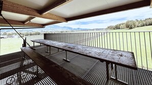 Un espace extérieur abrité, doté d’une table de pique-nique et d’un barbecue en métal, qui offre une vue sur un champ verdoyant et des montagnes lointaines.