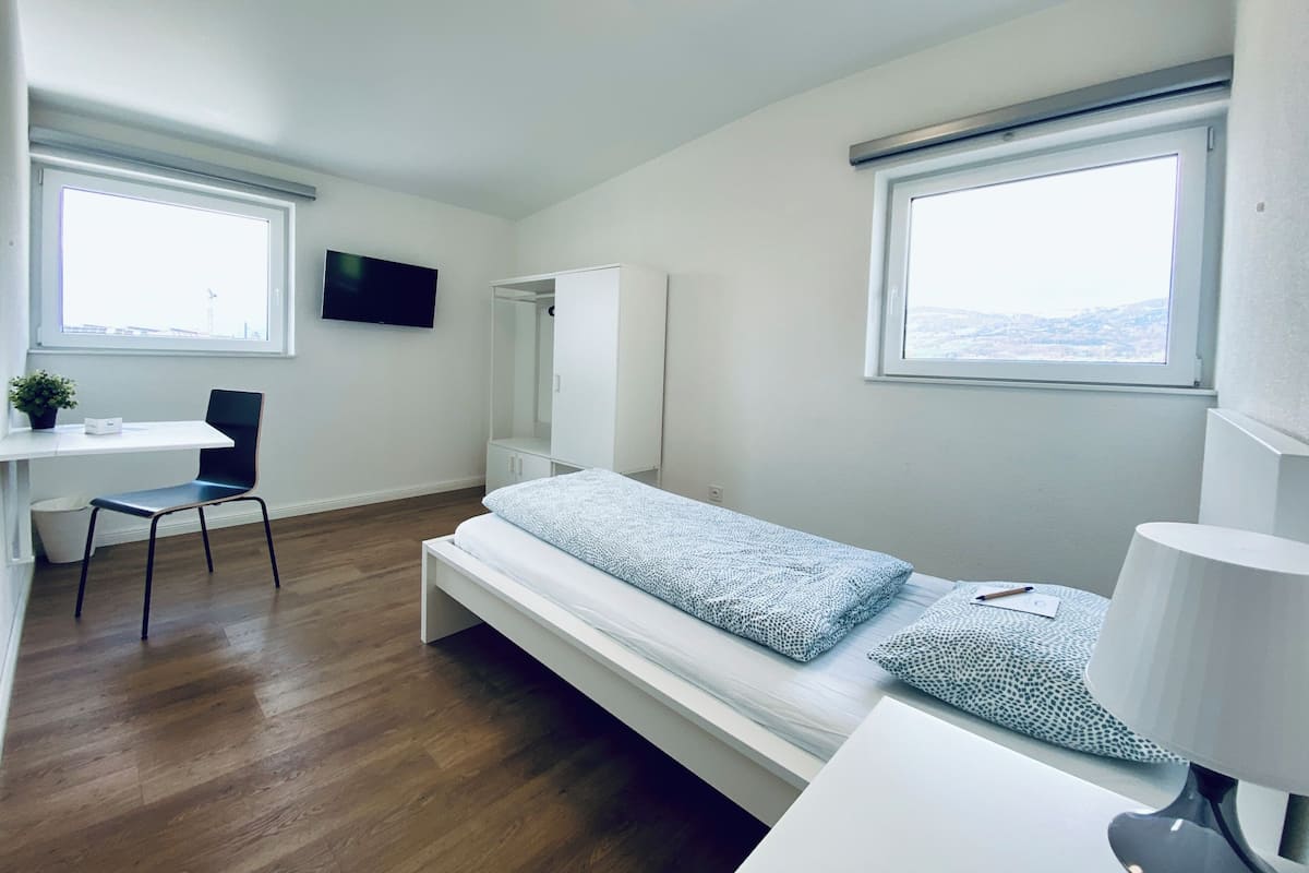 Une petite pièce propre, avec un lit, un bureau, une chaise, un téléviseur et une fenêtre.