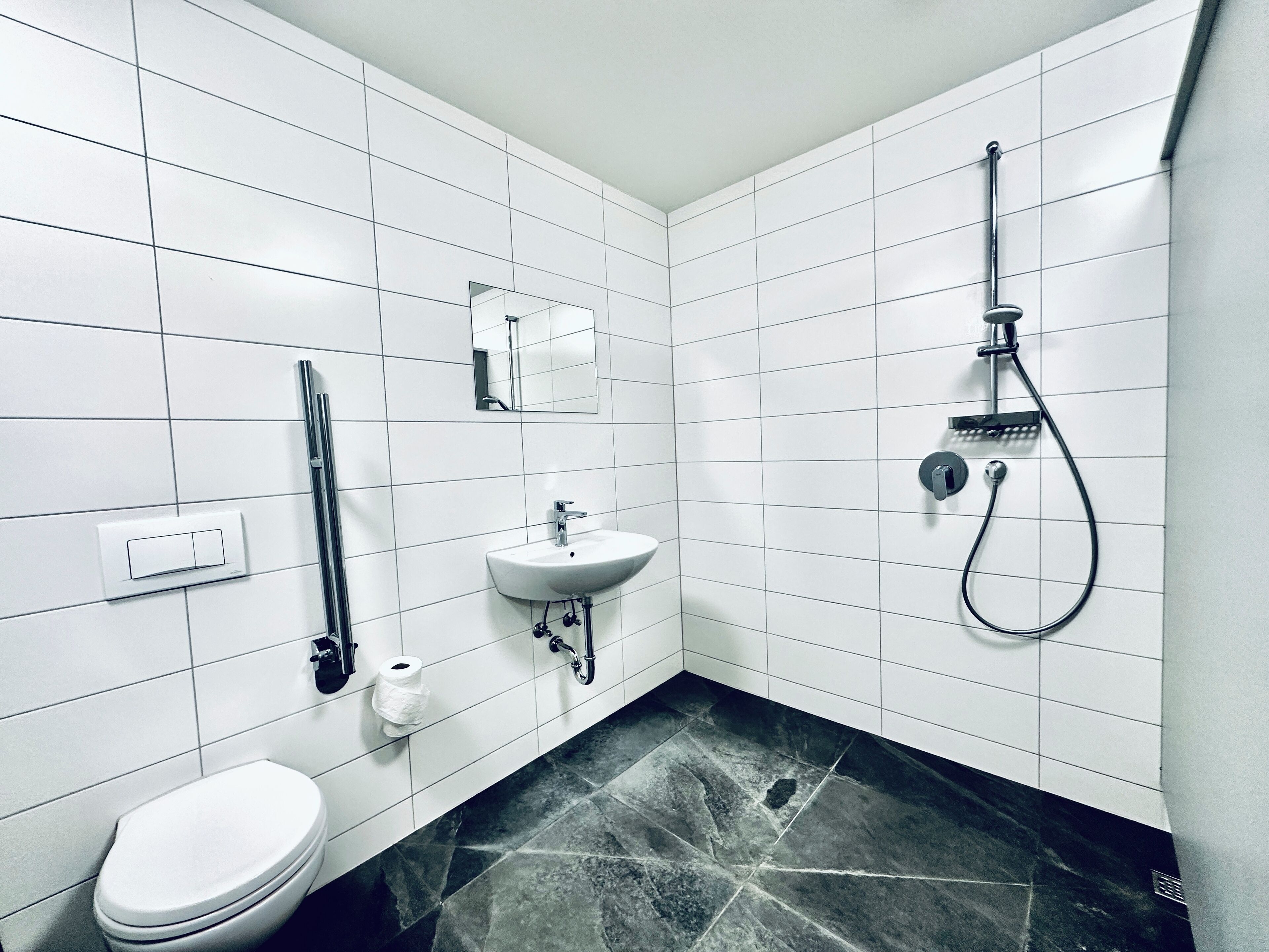A modern bathroom with white tiled walls, a black marble floor, a white toilet, a sink with a mirror, and a shower with a handheld showerhead.