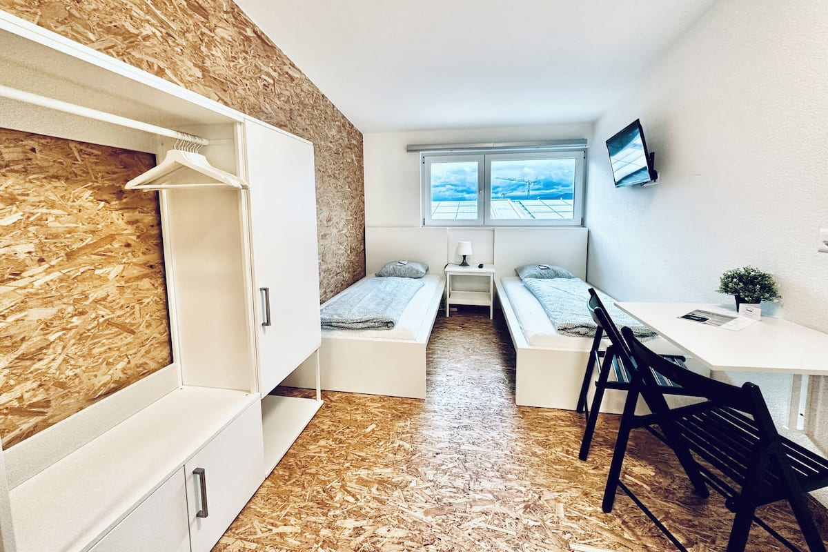 Une petite pièce, de taille modeste, comprenant un lit superposé, un bureau et une chaise. Une fenêtre offre une vue sur le ciel, et un téléviseur est fixé au mur.