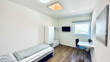 Una habitación pequeña y moderna con piso de madera, una cama con ropa de cama estampada, un escritorio con una silla, un televisor y una ventana con vistas.