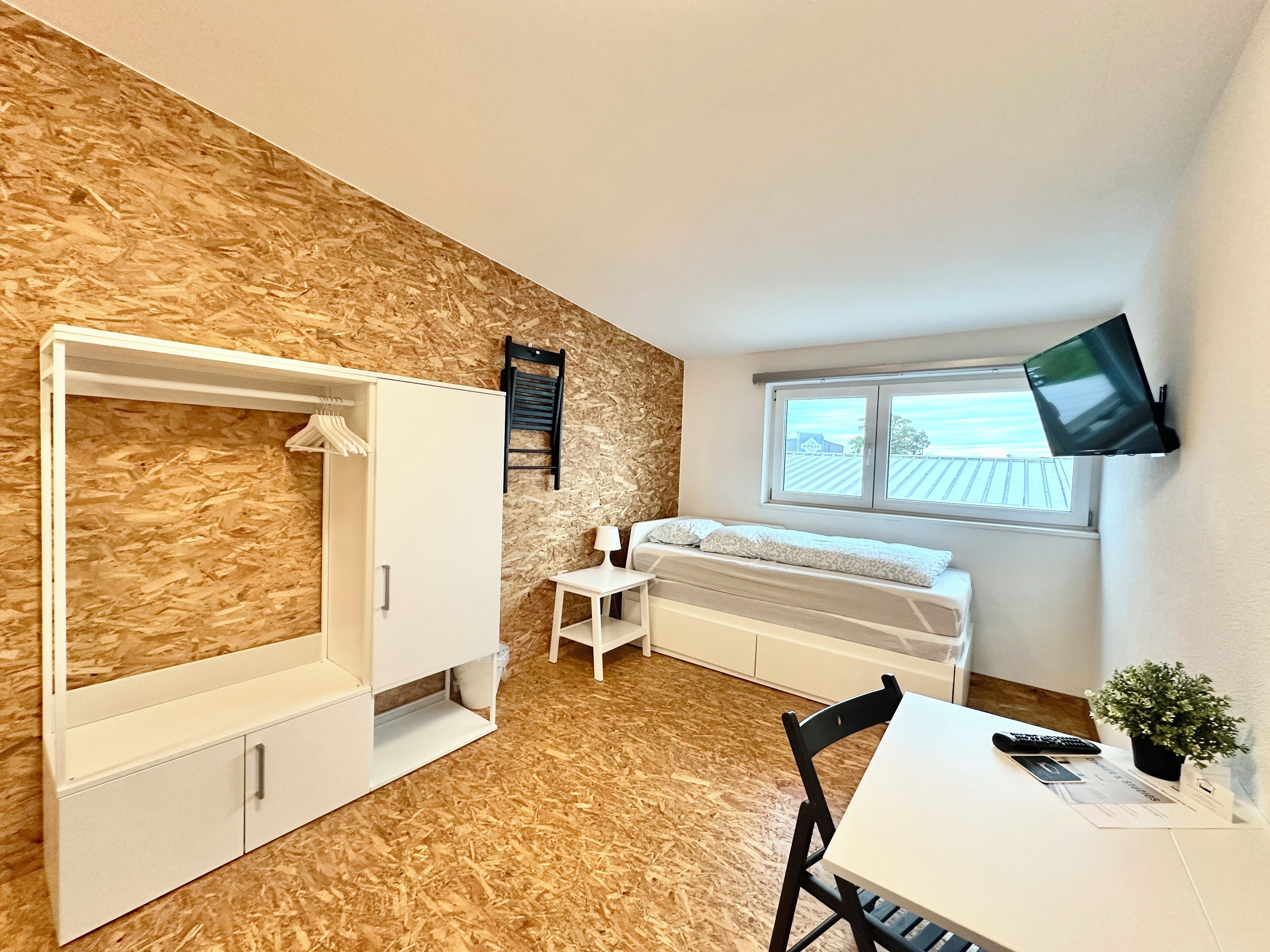 Een compacte kamer met een stapelbed, een bureau met stoel, een klein tafeltje, een aan de muur bevestigde televisie en een raam.